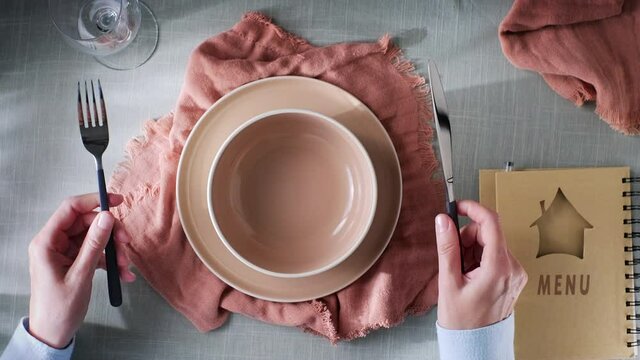 a restaurant visitor is waiting for food to be brought to him. There is a menu, plates and a knife with a fork on the table. also on the table are two rag napkins