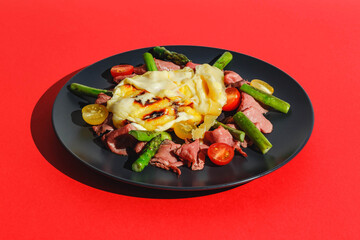 Delicious raclette cheese served over Roast Beef with Tomatoes and Asparagus, dish on red background. Hard light, deep shadow