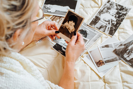 Close-up of middle-aged woman looking at old retro photograph of youth while relaxing on bed in bedroom, top view. Selective focus on vintage pictures, photos from family album. Nostalgia and memories