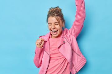 Upbeat European woman shakes arms celebrates victory makes triumph dance exclaims loudly closes eyes wears fashionable pink jacket isolated over blue background achieves goal or wis challenge - obrazy, fototapety, plakaty