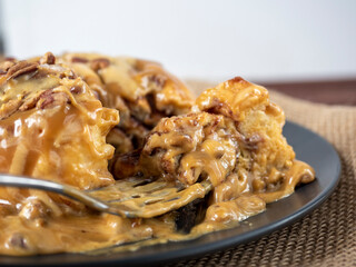 A close-up of a cinnamon bun sprinkled with caramel and decorated with nuts lies on a black plate and a linen napkin. Side view, a piece of dessert is taken with a fork. Selective focus
