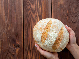Close-up of hands holding a round whole loaf of wheat bread. Brown wooden background, Top view, flat lay. Bakery concept. Copy space