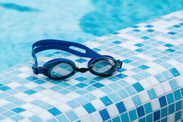 Blue professional swimming goggles with water drops on yellow lenses on a blue and white tiles of pool floor.