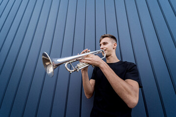 Young street musician playing the trumpet near the big blue wall - obrazy, fototapety, plakaty
