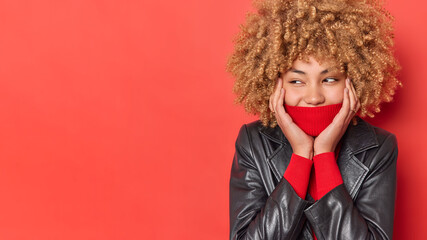 Studio shot of mysterious happy woman looks away covers mouth with collar of turtleneck keeps hands on face dressed in leather jacket isolated over red background blank copy space for promo.