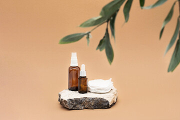 Blank amber glass essential oil bottle with pipette, glass spray bottle and reusable cotton sponges on natural stone podium. Organic spa cosmetic beauty product mock up.