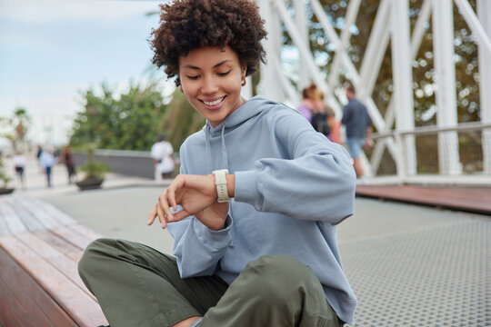 Outdoor shot of happy curly haired woman checks covered distance on smartwatch sits crossed legs against blurred background with people walking wears hoodie and trousers spends weekend at city