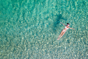 Top, aerial view. Athletic body of young beautiful woman in headphones listening music lying and swimming in white bikini in sea water on the sand beach. Drone, copter photo. Summer vacation.