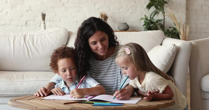 Latina woman and diverse kids enjoy hobby sit at table in living room holds pencils drawing pictures in paper album, spend leisure at home. Babysitting, daycare, childhood pastime and adoption concept