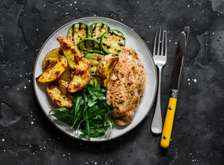 Delicious lunch - baked potatoes, chicken breast, grilled zucchini and arugula spinach salad on a dark background, top view i