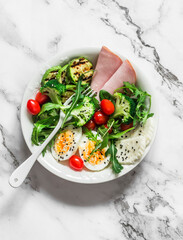 Delicious balanced breakfast, snack bowl - ham, boiled egg, grilled zucchini, green salad in one bowl on a light background, top view
