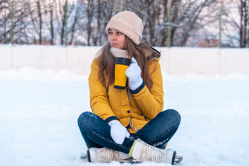 Fototapeta na wymiar Portrait of young woman in ice skates sitting on the snow, drinking coffee and looking away