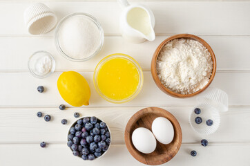 Recipe step by step. Ingredients for making lemon muffins with blueberries and a shtreisel on top. Butter, sugar, flour, eggs, milk, lemon zest on a white wooden background. Top view.