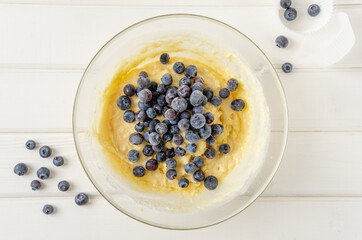Muffin dough with lemon zest and blueberries in a bowl on a white wooden background. Recipe step by step. Top view.