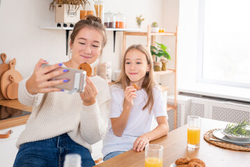 Obraz na płótnie Canvas Image of two happy girls sisters at kitchen indoors eat have a breakfast together take a selfie by phone.