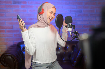 Young smiling muslim female singer wearing headphones with a microphone while recording song in a music studio with colorful lights.