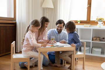 Young parents and little adorable son and daughter drawing together in sketchbook with coloured pencils sit at table in modern nursery, loving family enjoy leisure at home. Pastime and hobby concept