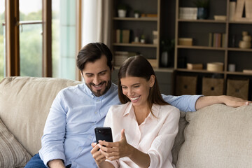 Young couple sits on sofa at home smiles looking at cellphone screen, spouses having pleasant conversation with family or friends living abroad using video call application. Modern tech, fun concept