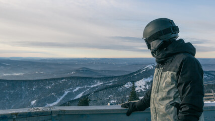 A snowboarder at an observation point in a ski resort leans on the railing and looks at a beautiful...