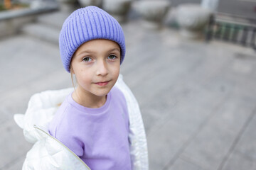 Cute little caucasian girl eight years old with blonde hair smiling outdoor. Kid wearing stylish shirt and knitted hat violet color. Trendy color of the 2022 year very peri