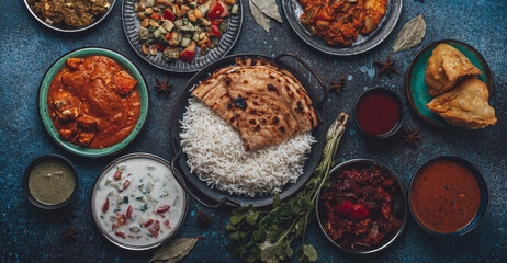 Assorted Indian ethnic food buffet on rustic concrete table from above: curry, fried samosa, rice biryani, dal, paneer, chapatti, naan, chicken tikka masala, mango lassi, dishes of India for dinner