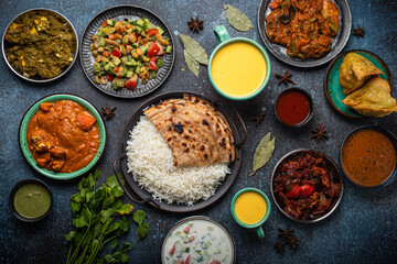 Assorted Indian ethnic food buffet on rustic concrete table from above: curry, fried samosa, rice biryani, dal, paneer, chapatti, naan, chicken tikka masala, mango lassi, dishes of India for dinner