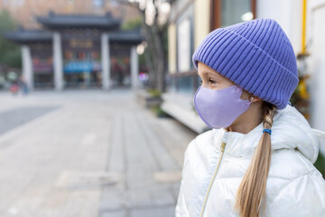 Cute little caucasian girl eight years old with blonde hair and protective face mask walking outdoor. Kid wearing stylish shirt and knitted hat violet color. Trendy color of the 2022 year very peri
