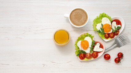 Nicely served breakfast on Valentine's Day. Two heart-shaped plates with fried eggs, tomatoes and cheese, coffee and juice on a white wooden table. View from above. Banner. Healthy breakfast. Banner