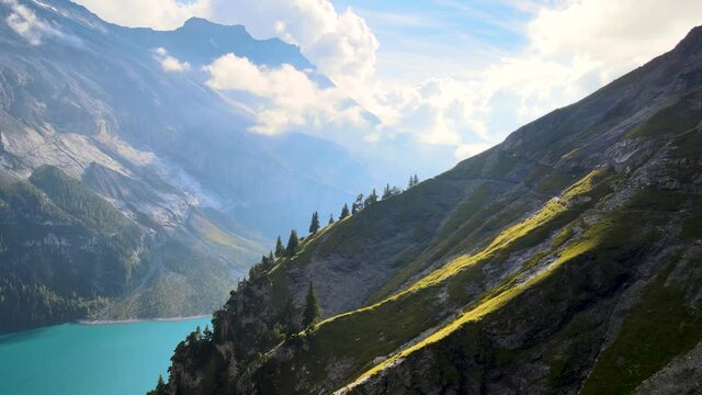 Aerial drone shot of the side of a rocky mountain on a sunny day. Walking path in the middle of the swiss alps on a green cliff with Blue glacier lake Oeschinen in the background
