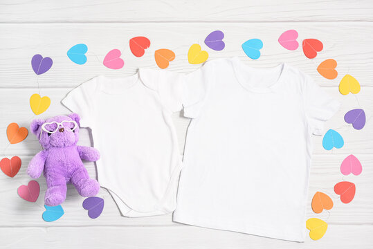 Mockup white child shirt and newborn onesie with paper colorful hearts and teddy bear in glass. Happy Valentines Day baby bodysuit apparel flatlay on white wood background, Top view, copy space.
