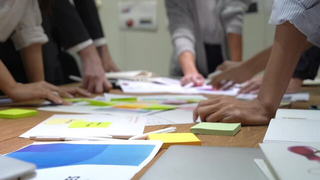 Selective focus of a group of business people's hands picking up paper sheet with sticky notes that scattered on a table to see an information while brainstorming for business plan during a meeting.
