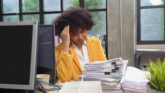 Unhappy Asian female employee with an Afro hairstyle, sitting at desk behind computer and a large stack of paperwork resting head on hand and looking forward with a boring face, overworked office lady