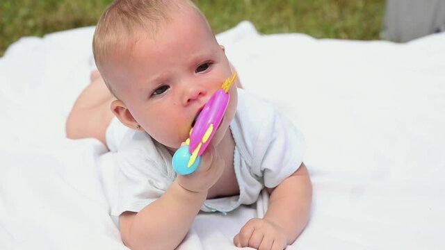 Little newborn girl lying on tummy chewing rattle on blanket, summer day outdoor. Infant having fun outdoors. Infant baby child resting playing learning to crawl. Motherhood teething, babyhood concept