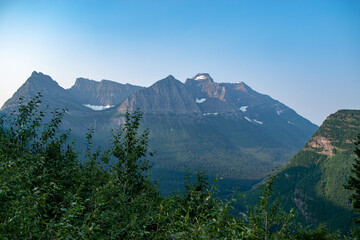 Snowy and rocky mountains seen from forest of green trees with blue sky in Glacier National Park