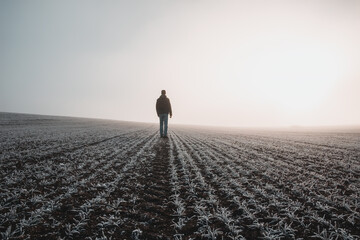 Young caucasian man in jacket standing on meadow, field covered with hoarfrost in misty fog at...