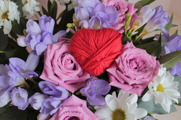 Against the background of pink roses, blue freesias and white chamomile lies a red heart made of threads.