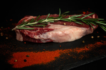 Raw marbled beef ribeye steak with rosemary branch and spices. A piece of meat with fatty veins on a stone black board. Side view.