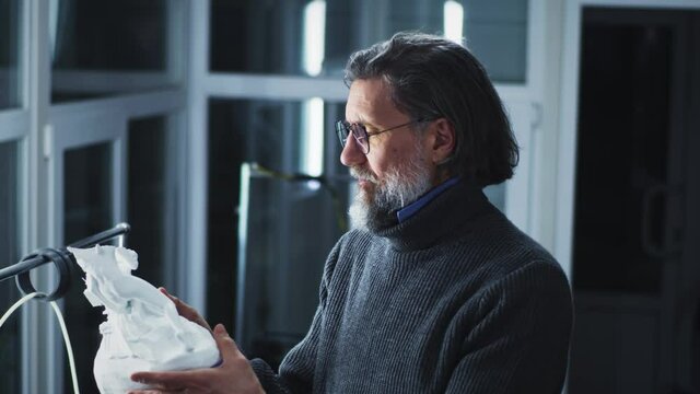 Tilt up view of mature man in sweater adjusting glasses and inspecting plastic statuette while working in 3D printing studio