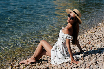 A beautiful young woman in a hat, glasses and a light dress is sitting with her back to the ocean against the background of huge rocks on a sunny day. Tourism and tourist trips.