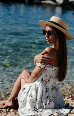 A beautiful young woman in a hat, glasses and a light dress is sitting with her back to the ocean against the background of huge rocks on a sunny day. Tourism and tourist trips.