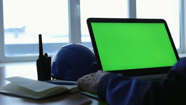 Male Engineer work at Laptop Computer with Green Screen inside office. View from back with hard hat and walkie-talkie at background. Great for Mockup usage.