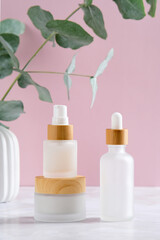 Elegant cosmetics containers and eucalyptus in vase on pink background. Essential oil bottles and cream jar. Skincare beauty products packaging design