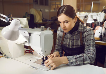 Serious concentrated young caucasian female shoemaker in uniform working. Woman worker sewing on machine at workplace. Footwear factory, fashion industry, handmade boots production concept