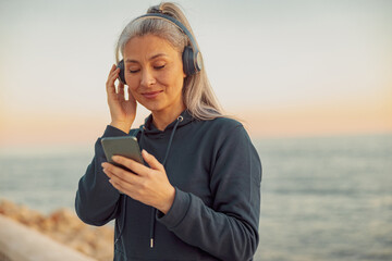 Portrait of happy lady in black hoodie listening to music in headphones, standing on background of sea holding and looking on smartphone