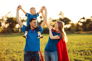 Fototapeta premium Mother, father and son in superhero costumes. Parents hold their son in their arms and play superhero outdoors