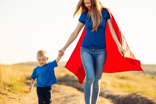 Super Mom And Her Son Walk Forward Holding Hands. Cheerful Family, A Woman In A Red Raincoat As A Superhero