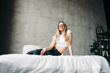 Young attractive caucasian woman in eyeglasses with blonde hair sitting on a bed home.