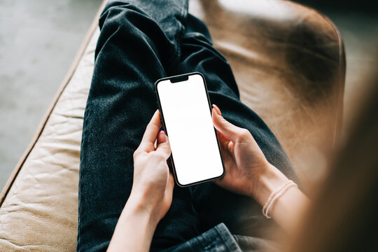 Woman holding mobile phone with white screen mock up, resting on a sofa in living room at home.	