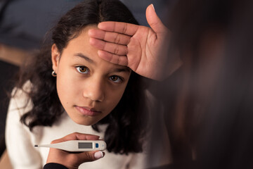 Girl looking at camera while her mother check the temperature with a thermometer at home.