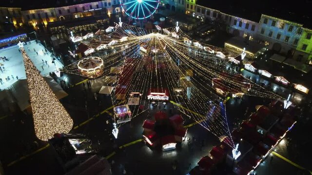 Aerial drone view of The Big Square in Sibiu at night, Romania. Old city centre decorated for Christmas. Ferris wheel, skating rink, old traditional buildings, people